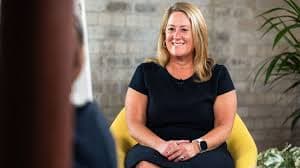 Smiling blonde woman in a navy dress sits in a yellow chair during an interview.