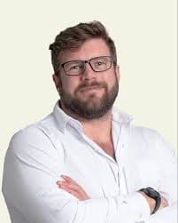 Bearded man with glasses wearing a white shirt, posing with arms crossed against a neutral background.